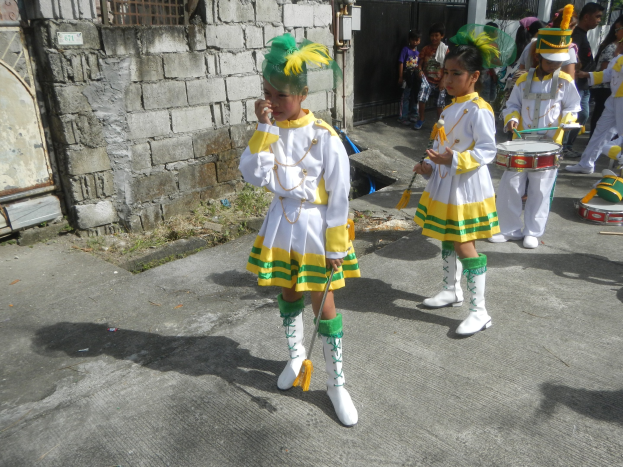 Gruppe junger Mädchen in weißen und gelben Outfits, die marschieren und Musikinstrumente spielen, vorbei an einer Wand, einem Tor und Bäumen.