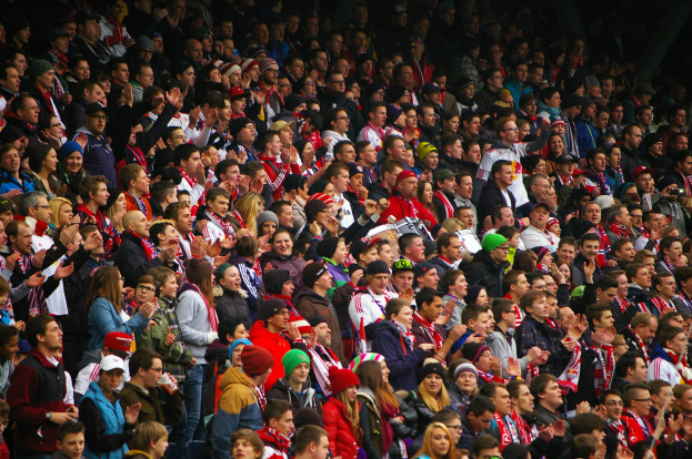 Eine große Menge in einem schwach beleuchteten Stadion hält die Hände hoch, einige tragen Mützen und Schals und erzeugen eine dramatische Atmosphäre.