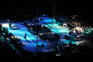 Eine Gruppe von Menschen, die auf einer Bühne mit Lautsprechern, Scheinwerfern und Mikrofonen performs, umgeben von einer großen Menschenmenge im dunklen Hintergrund.