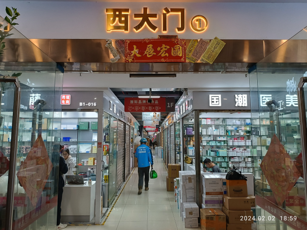 Innenansicht einer chinesischen Apotheke mit Glaswänden, Regalen voller Artikel, verstreuten Kartons auf dem Boden, einer Pflanze links und einer Namensschilder oben.
