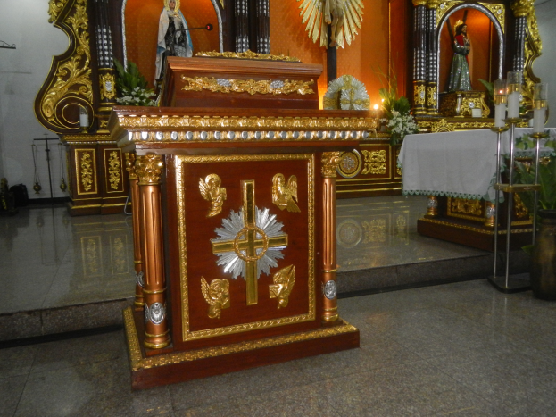 Ein Altar in einer Kirche mit einem Kreuz, Statuen, Blumenbouquets, Kerzen mit Ständern, einem Tisch mit einer Decke und anderen Gegenständen, während die Wände mit Lichtern geschmückt sind.