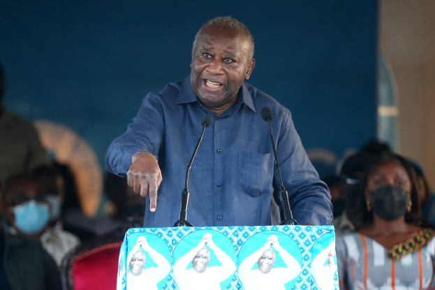 Ein Mann in einem blauen Hemd spricht in zwei Mikrofone an einem Rednerpult und spricht zu einer Menge, von der einige Masken tragen, mit einer Wand im Hintergrund.