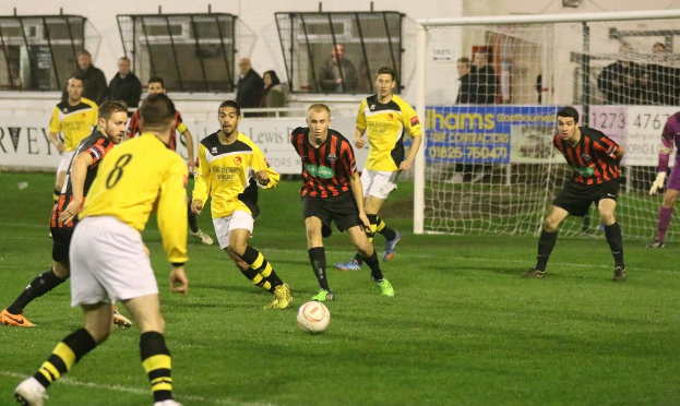 Ein Sportfeld mit mehreren Männern, die Fußball spielen, ein Netz auf der rechten Seite, eine Tafel mit Text im Hintergrund und Zuschauer, die hinter der Tafel stehen, sowie eine Wand und Fenster weiter im Hintergrund.