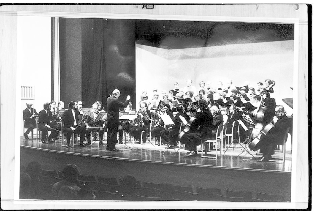 Ein Schwarz-Weiß-Foto eines Dirigenten in Anzug und Krawatte, der auf einer Bühne steht und ein Orchester mit Geigen, Celli und Flöten leitet, während ein Publikum in Stühlen aufmerksam zuhört.