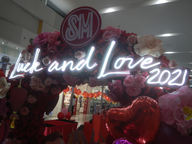 Ein Geschäft mit einem "Glück und Liebe 2021"-Schild, umgeben von Blumen, Ballons und Dekorationen, mit anderen Geschäften, Lichtern und einer Wand im Hintergrund, die eine festliche Stimmung für eine Valentinstagsfeier schaffen.