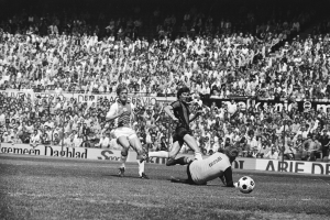 Schwarzes Foto von Manner, die auf einem Feld Fussball spielen, mit Zuschauern im Hintergrund und einem Banner mit der Aufschrift "Bundesliga 1978-79 Ajax 1-1 Hannover 96."