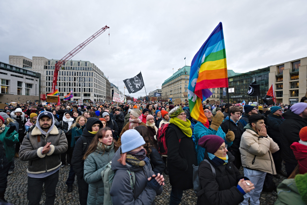 Eine große Gruppe von Menschen bei einer LGBTQ+-Rechtsdemo in Berlin, die Fahnen und Plakate schwenken, mit Gebäuden, einem Kran und einem bewölkten Himmel im Hintergrund.