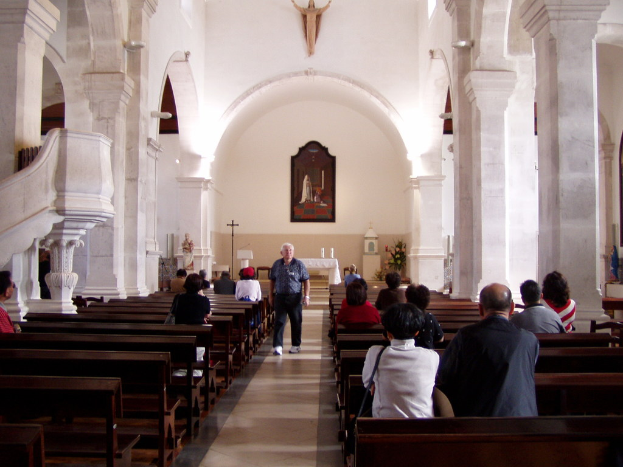 Ein älterer Mann geht den Mittelgang einer Kirche entlang, flankiert von Sitzreihen mit sitzenden Personen, während Fotos und Idole die Wände im Hintergrund schmücken.