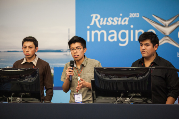 Drei Jungen stehen neben einem Schreibtisch mit zwei Computern, einer spricht in ein Mikrofon und trägt ID-Karten mit roten Markierungen, vor einer Wand mit einer Werbetafel, auf der "Russia Imagine 2013" steht.