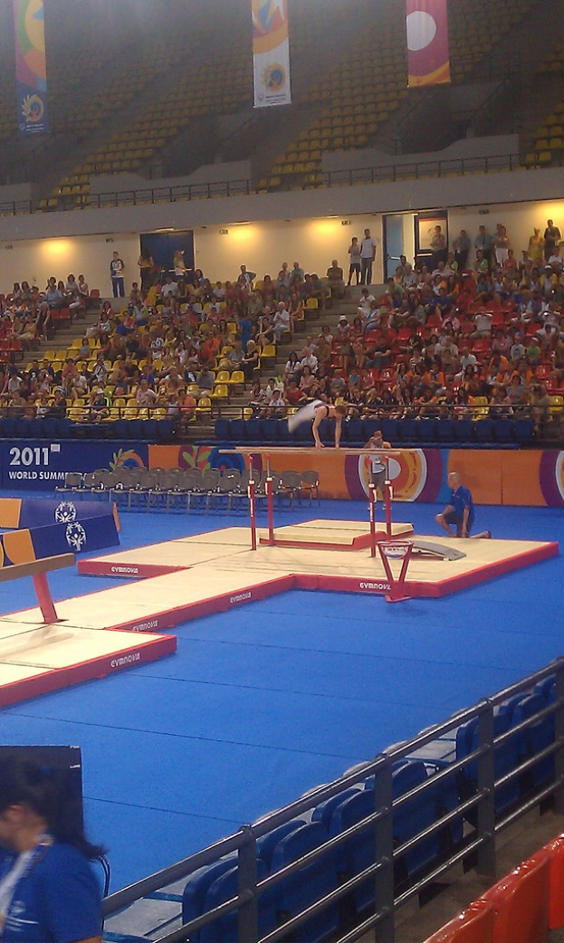 Eine Stadionszene mit Menschen auf Stühlen, andere im Hintergrund, Werbetafeln oben, Treppe in der Mitte und eine Person, die Gymnastik macht.