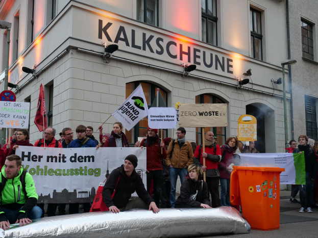 Eine Gruppe von Menschen mit Protestschildern und Plakaten vor einem Gebäude, mit zwei Personen im Vordergrund und einem Müllcontainer auf der rechten Seite, vor einem Hintergrund von städtischen Gebäuden mit Fenstern und Schildern in Deutschland.