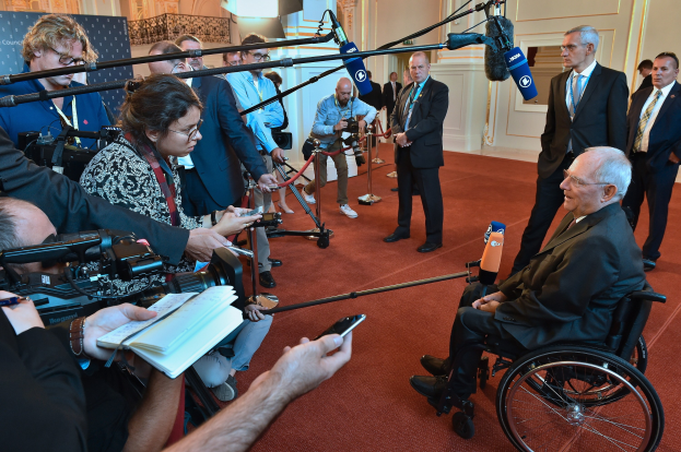 Ein Mann im Rollstuhl in Anzug und Krawatte umgeben von Reportern mit Kameras und Mikrofonen sowie einem Banner und einer Wand im Hintergrund.