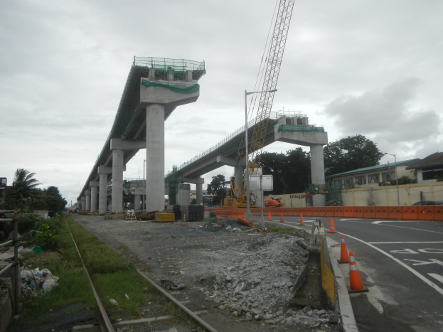 Baustelle mit einer Brücke im Hintergrund, eine Straße mit Absperrbaken rechts, Steine und Gras am Boden, eine Bahnschiene links, Bäume und Gebäude auf beiden Seiten der Straße und ein bewölkter Himmel.