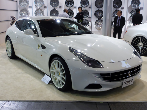 Ein weißer Ferrari FF auf einer Autauschau umgeben von Menschen in verschiedenen farbigen Kleidern, mit mehreren Rädern an der Wand im Hintergrund.