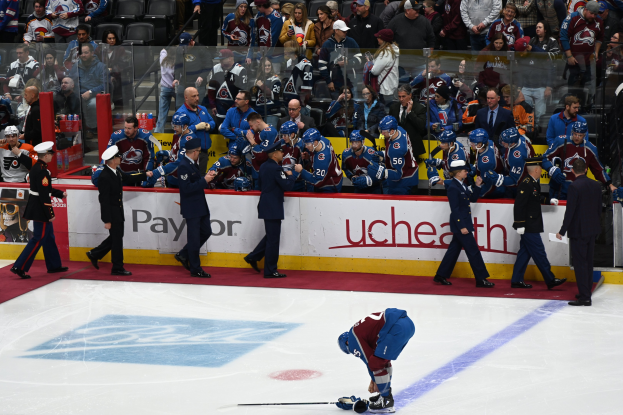 Ein Eishockeyspieler auf dem Eis umgeben von Teamkollegen und Gegnern, mit Zuschauern und Arena-Schildern im Hintergrund.