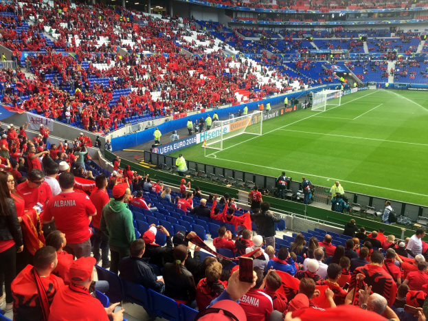 Eine große Menge Menschen in einem hell erleuchteten Stadion schaut einem Fußballspiel zu, mit Sitzplätzen und Stehplätzen, Schildern, einem Tor und Deckenbeleuchtung.