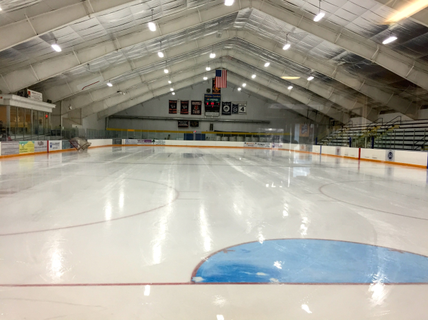 Große Indoor-Eisbahn mit Deckenbeleuchtung, Werbetafeln, einer Rückwand, Treppen mit Geländern und einigen Menschen, was auf eine Sportanlage hinweist, die likely für Hockey genutzt wird.