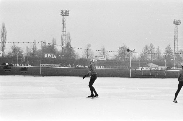 Zwei Menschen, die auf einer Eisbahn im Schnee Schlittschuh laufen, umgeben von einem Zaun, Bannern, Pfosten, Lichtern, Bäumen und einem klaren Himmel, dargestellt in Schwarz-Weiß.