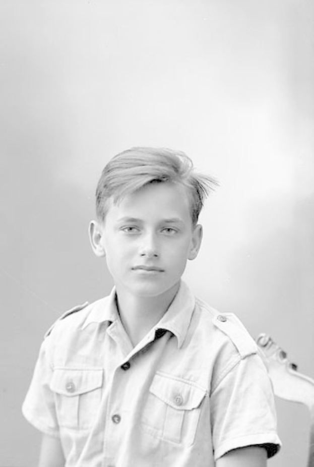 Ein junger Junge in einem Schwarz-Weiß-Foto steht mit einer Gitarre in der Hand, trägt ein Hemd mit ernstem Gesichtsausdruck, kurze dunkle Haare und konzentrierte Augen auf dem Instrument.